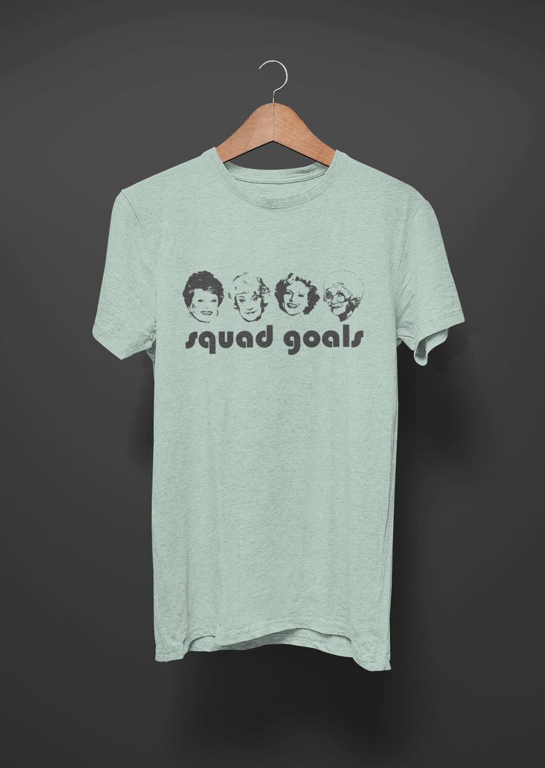 May include: A pale green t-shirt featuring a black graphic of four women's faces and the words "squad goals" printed underneath. The t-shirt is suspended on a wooden hanger against a dark grey backdrop.