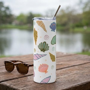 Puede incluir: Un vaso de acero inoxidable blanco con un colorido diseño de conchas marinas. El diseño incluye varias ilustraciones de conchas marinas en tonos rosa, azul, verde y dorado. Una pajita plateada está insertada en la tapa. Gafas de sol sobre una superficie de madera.