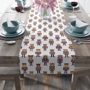 May include: A white table runner featuring a repeating pattern of colorful nutcracker figures. The nutcrackers are in various colors, including purple, gold, green, and black, with crowns and regal attire. The runner is set on a wooden table with place settings.
