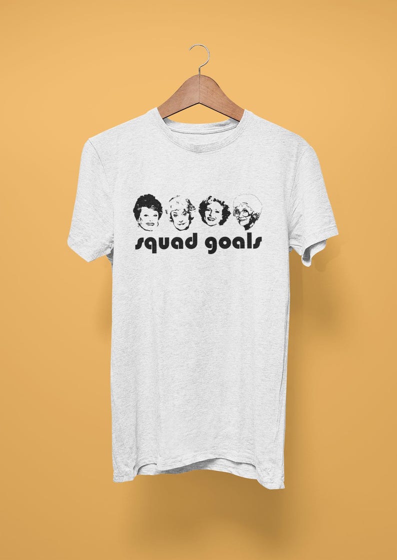 May include: A light grey t-shirt with a black and white graphic of four women's faces and the words "squad goals" in black. The t-shirt is displayed on a wooden hanger against a yellow background.