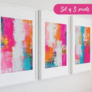 May include: Set of three abstract art prints in white frames. The prints feature bold strokes of pink, orange, yellow, and blue paint.