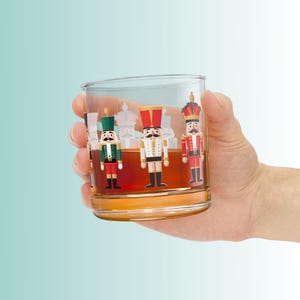 May include: A clear glass tumbler with a festive design featuring three toy soldier figures in red, green, and white. The glass is filled with a brown liquid and held by a hand against a light blue background.