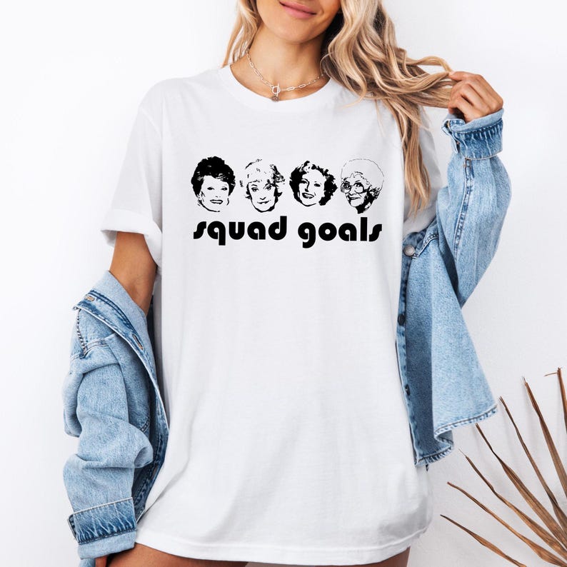 May include: White t-shirt with a black and white graphic of four faces and the words "squad goals". The t-shirt is a casual, unisex style.