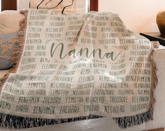 May include: A cream-colored woven throw blanket featuring a repeating pattern of names including Eleanor, Theodore, Nicholas, Timothy, Zachary, Benjamin, Remy, and Sarah, with the word Nanna printed in large, dark green cursive script.