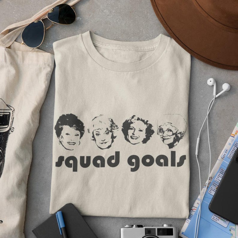 May include: A cream-coloured t-shirt with four black and white portraits of women and the words "squad goals" in a bold, sans-serif font. The shirt is folded on a light grey surface.