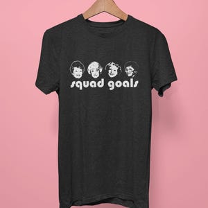 May include: Dark grey t-shirt with a white graphic of four women's faces and the words "squad goals." The shirt hangs on a wooden hanger against a pink background. The t-shirt is made of a soft material.