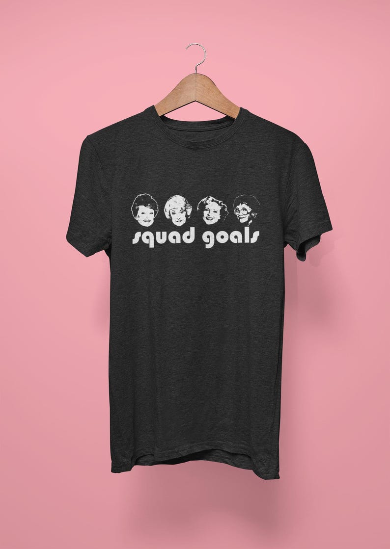 May include: Dark grey t-shirt with a white graphic of four women's faces and the words "squad goals." The shirt hangs on a wooden hanger against a pink background. The t-shirt is made of a soft material.