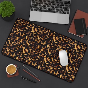 May include: A rectangular desk mat with a tortoiseshell pattern in shades of brown and gold. A white computer mouse sits on the mat, with a laptop, notebooks, pens, and a coffee cup nearby.