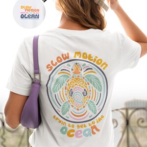 Puede incluir: Camiseta blanca con un gráfico colorido de una tortuga y el texto "slow motion tryin to get to the ocean" en la espalda.