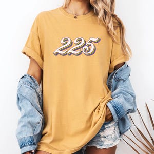 Puede incluir: Una camiseta amarilla mostaza con un gráfico retro blanco, morado y amarillo del número 225.