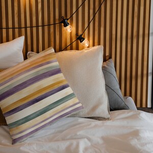 May include: A white bed with a striped pillow, a beige pillow, a brown corduroy pillow, and a white pillow. The bed is against a wood wall with string lights.
