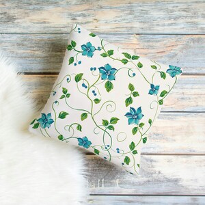 May include: Square decorative pillow with a white background and a floral pattern. The design features teal blue flowers, green leaves, and vines. The pillow is placed on a white fluffy surface and a wooden background.