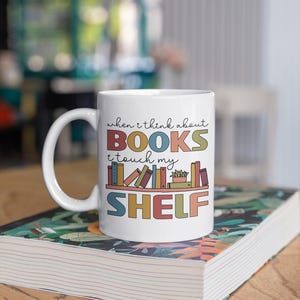May include: White ceramic mug with a colorful graphic that reads "When I think about books I touch my shelf". The graphic features a bookshelf with colorful books and a small potted plant.