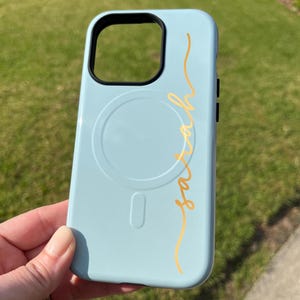 Pu&ograve; includere: Custodia per telefono azzurra con un ritaglio nero per la fotocamera e il nome "Sarah" in oro in corsivo. La custodia presenta un design circolare e un design rettangolare sul retro. Su sfondo verde.