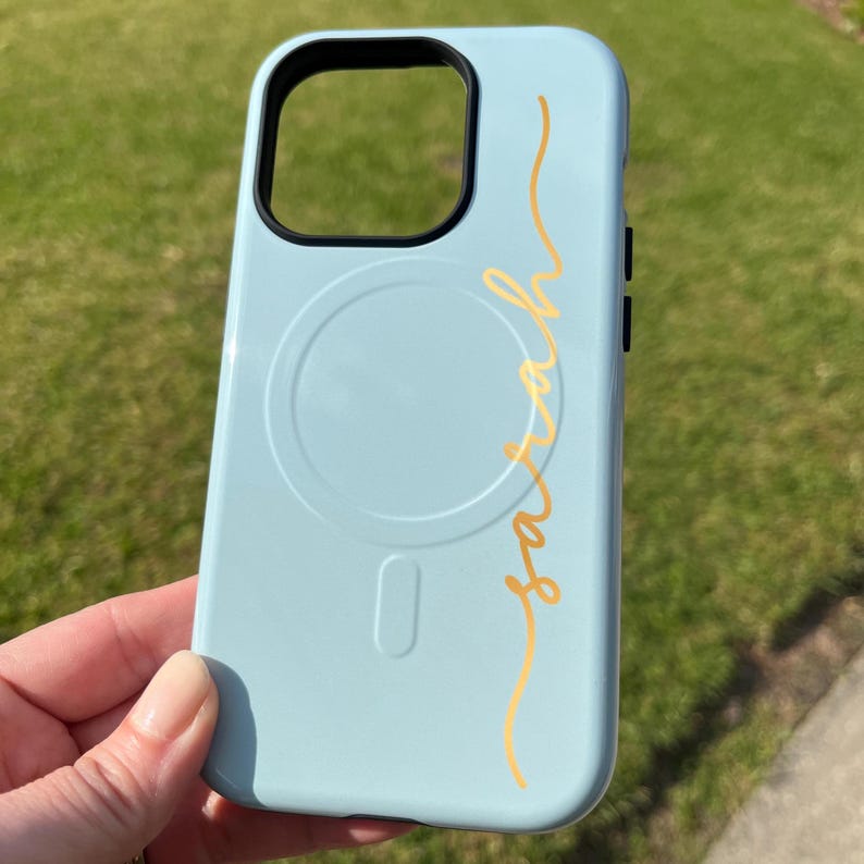 Pu&ograve; includere: Custodia per telefono azzurra con un ritaglio nero per la fotocamera e il nome "Sarah" in oro in corsivo. La custodia presenta un design circolare e un design rettangolare sul retro. Su sfondo verde.