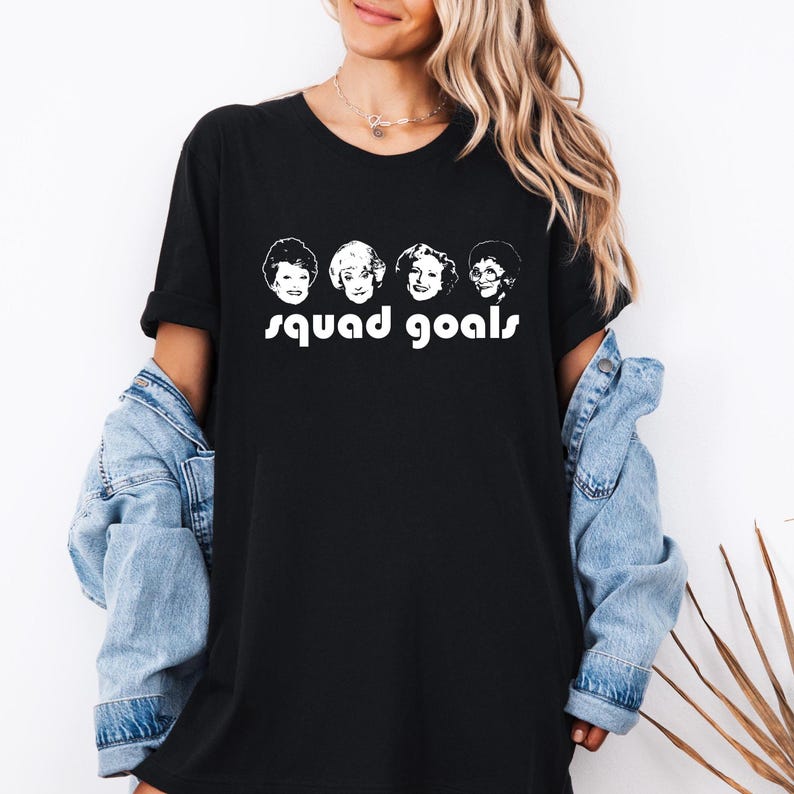 May include: Black t-shirt with a white graphic of four women's faces and the words "squad goals". The t-shirt is a classic crew neck style, suitable for casual wear. The design is simple but eye-catching, making it a versatile wardrobe staple.