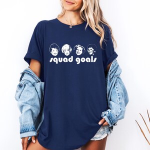 May include: Navy blue t-shirt with a white graphic of four faces and the words "squad goals". The top has short sleeves and a loose fit. The design is a simple, bold statement.