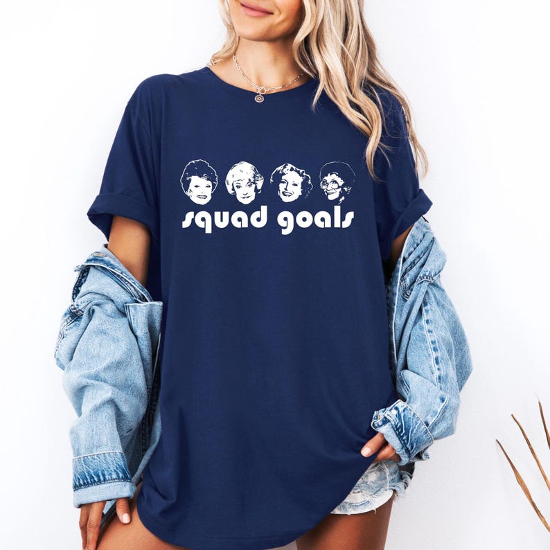 May include: Navy blue t-shirt with a white graphic of four faces and the words "squad goals". The top has short sleeves and a loose fit. The design is a simple, bold statement.