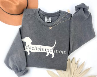 Puede incluir: Una sudadera gris con la frase "dachshund mom" y una silueta blanca de perro salchicha. También se ven un sombrero marrón y un collar dorado. La sudadera es de cuello redondo.