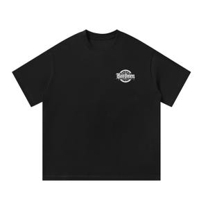 May include: A black short-sleeved t-shirt featuring a small, circular white graphic on the upper left chest. The graphic contains the text "Bad Omen" in a stylised, gothic-inspired font, surrounded by a thin circular border and decorative elements.