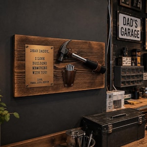 May include: A rectangular wooden wall plaque featuring a brass plate with the engraved text: DEAR DADDY, I LOVE BUILDING MEMORIES WITH YOU! LOVE, EMMA, JOSH, KEVIN. A metal hammer is mounted above a small brown leather pouch containing silver nails.