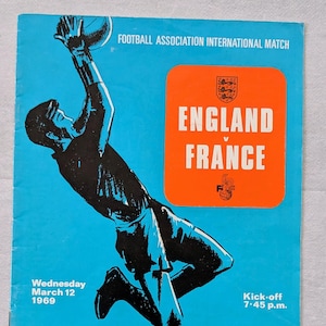 May include: Vintage football program cover featuring a player reaching for a ball. The cover is blue with an orange rectangle displaying "England v France." Text includes "Football Association International Match" and "Official Programme."