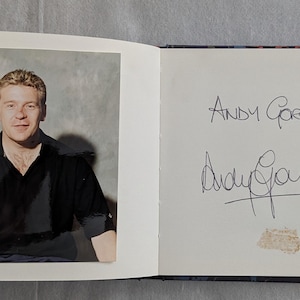 May include: A photograph of a man in a black polo shirt with a small logo on the chest. The image is in a book, with the man's name and signature on the facing page. The background is a light gray.
