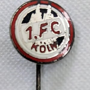 Puede incluir: Un pin redondo con fondo blanco y borde rojo. El pin presenta el texto "1. FC KOLN" en letras rojas. El pin tiene un alfiler de metal plateado en la parte posterior.