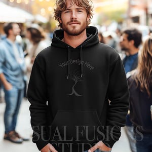 May include: Black hooded sweatshirt with a front pocket and drawstring hood. The sweatshirt features the text "Your Design Here" above a tree graphic and the text "SUAL FUSIONS STUDIO" at the bottom. The model is wearing blue jeans.