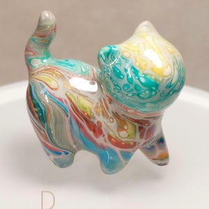 May include: A small glass cat figurine featuring a marbled swirl pattern in shades of turquoise, yellow, orange, and white. The cat is standing with its tail held upright, and the letter D is printed in a light tan colour below the figurine.