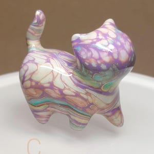 May include: A small, decorative cat figurine with a marbled design. The cat is primarily white, purple, and teal, with a glossy finish. The figurine has a raised tail and is standing on all fours. The letter "C" is visible on the base.