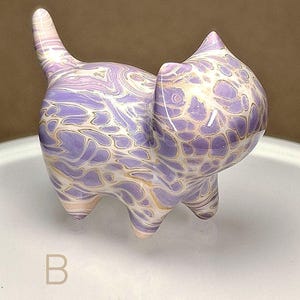 May include: A small, ceramic cat figurine with a purple and white marbled design. The cat is standing on a white surface with the letter "B" printed on it.