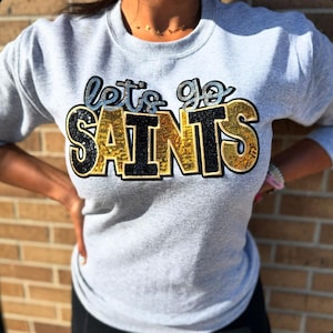 May include: A gray sweatshirt with the phrase "let's go SAINTS" in a sequined design. The text features black, gold, and blue sequins. The sweatshirt is a crew neck style.