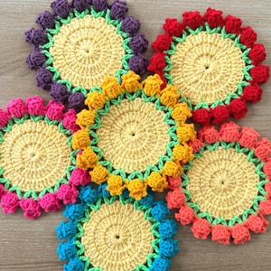May include: A collection of six handmade crochet coasters in a flower design. Each coaster features a yellow center, a green border, and a ring of colorful, three-dimensional petals in shades of purple, red, pink, orange, yellow, and blue.