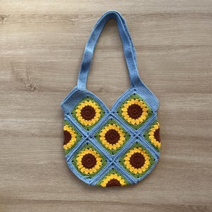 May include: A blue crocheted tote bag with a sunflower pattern. The bag has two straps and is made of a soft, textured yarn.