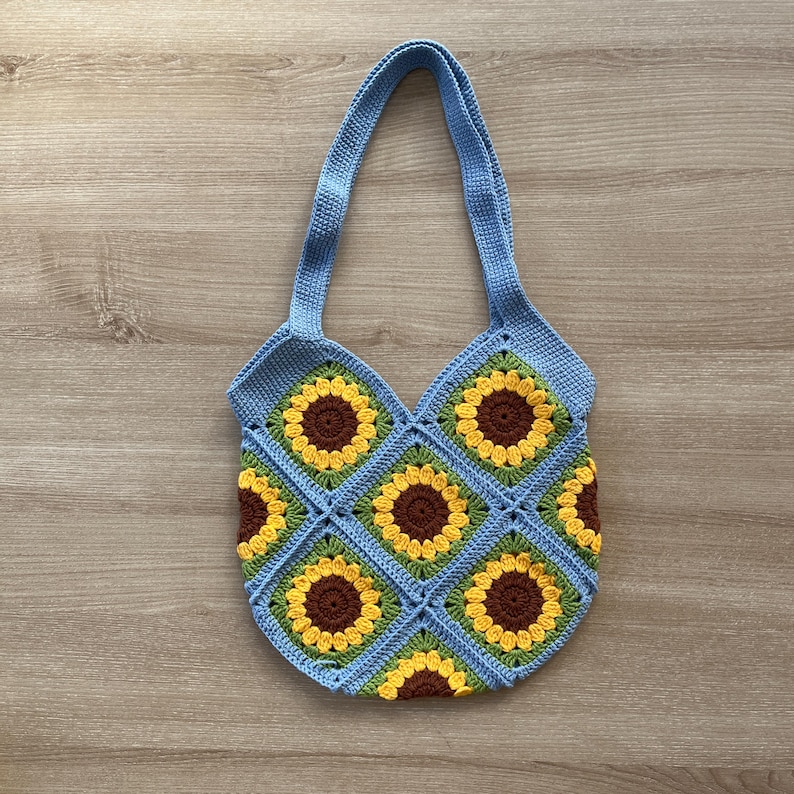 May include: A blue crocheted tote bag with a sunflower pattern. The bag has two straps and is made of a soft, textured yarn.