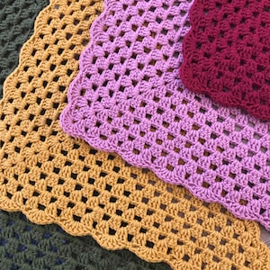 May include: Four crocheted squares in different colours: mustard yellow, purple, burgundy, and dark green. The squares are made with a granny stitch pattern.