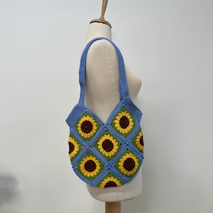 May include: A blue crocheted tote bag with a sunflower pattern. The bag has a single strap and is made of yarn.