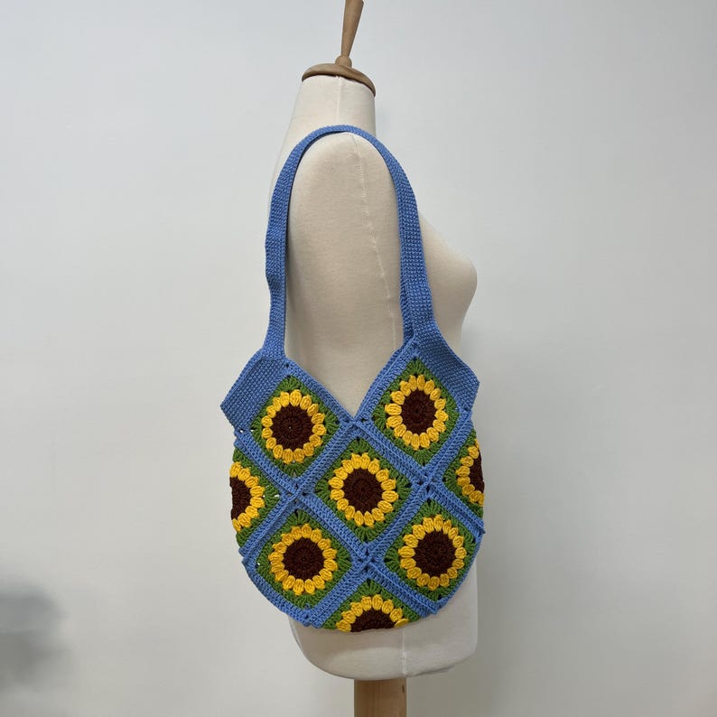 May include: A blue crocheted tote bag with a sunflower pattern. The bag has a single strap and is made of yarn.