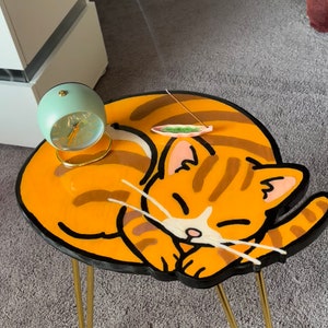 May include: A small orange and brown cat-shaped side table with gold metal legs. The table top is painted to look like a sleeping cat with a black outline. A small green and white incense holder is on the table.