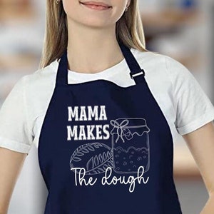 Può includere: Un grembiule blu navy con testo bianco che recita "Mama Makes The Dough". Il grembiule ha una tasca sul davanti e un'illustrazione bianca di un barattolo di marmellata e una pagnotta di pane.