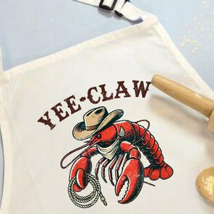 May include: White apron with a red lobster wearing a cowboy hat and the text "Yee-Claw".
