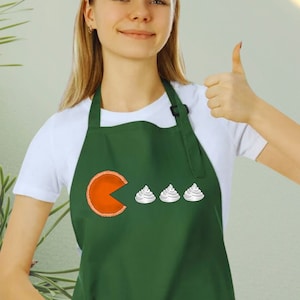 May include: A woman wearing a green apron with a white design of a slice of pie being eaten by three scoops of ice cream. The text "Pac-Pie" is embroidered on the apron.