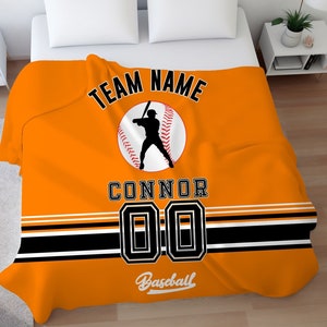 May include: Orange baseball-themed fleece blanket with black and white stripes. The blanket features a baseball player silhouette, a baseball, the text "TEAM NAME", "CONNOR", "00", and "Baseball".