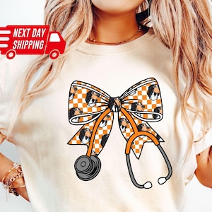 May include: Ivory-colored t-shirt featuring a large bow design with a checkered pattern in orange and white, and a stethoscope graphic. The bow design includes a repeating image of a dog's face. The shirt has short sleeves and a crew neck.