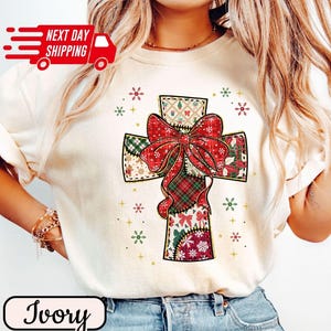 以下が含まれることがあります： アイボリー色のTシャツ。パッチワークの十字架のデザインで、大きな赤いリボンが付いています。十字架は、チェック柄や花柄など、さまざまな模様で装飾されており、雪の結晶で囲まれています。シャツには「Ivory」の文字も入っています。