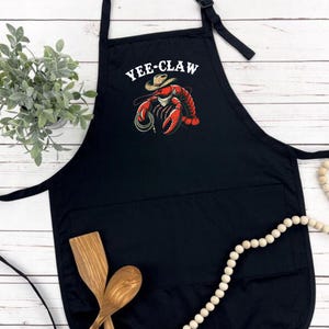 May include: Black kitchen apron with a pocket and a white graphic of a lobster wearing a cowboy hat with the text "Yee-Claw".