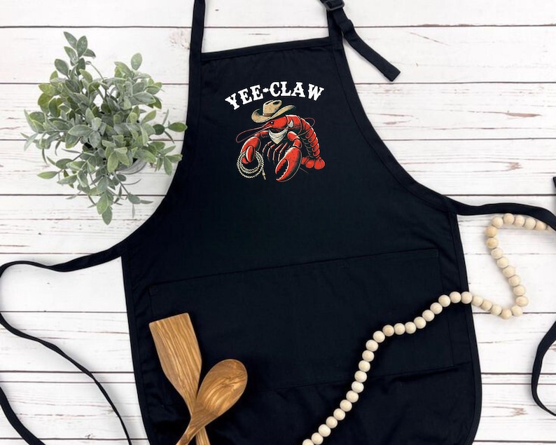 May include: Black kitchen apron with a pocket and a white graphic of a lobster wearing a cowboy hat with the text "Yee-Claw".