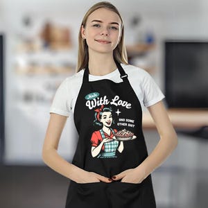 Puede incluir: Delantal negro con una ilustraci&oacute;n retro de una mujer sosteniendo un pastel. El delantal tiene el texto "Made With Love and some other shit". La mujer de la ilustraci&oacute;n lleva un pa&ntilde;uelo rojo y una camisa a cuadros rojos y azules.