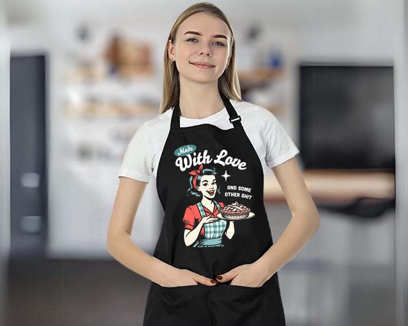 Puede incluir: Delantal negro con una ilustraci&oacute;n retro de una mujer sosteniendo un pastel. El delantal tiene el texto "Made With Love and some other shit". La mujer de la ilustraci&oacute;n lleva un pa&ntilde;uelo rojo y una camisa a cuadros rojos y azules.